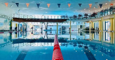 Blick in die leere Schwimmhalle, über das Schwimmerbecken im Phönix-Bad, der Mitte eine rote Trennleine zwischen den Schwimmbahnen; die Wasseroberfläche ist ganz still und die Umgebung spiegelt sich im Wasser