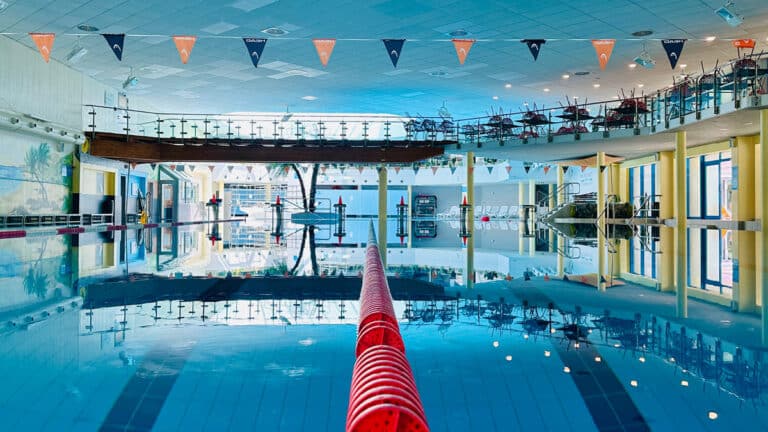 Blick in die leere Schwimmhalle, über das Schwimmerbecken im Phönix-Bad, der Mitte eine rote Trennleine zwischen den Schwimmbahnen; die Wasseroberfläche ist ganz still und die Umgebung spiegelt sich im Wasser